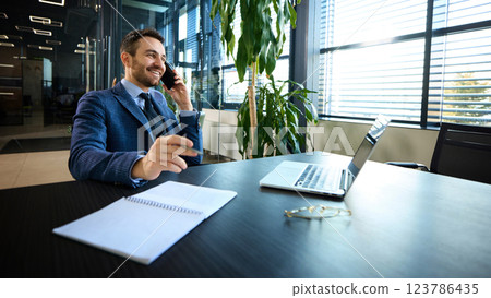 Cheerful businessman smiling during phone call, with laptop and office supplies on desk, under modern geometric ceiling lights Cheerful businessman smiling during phone call, with laptop and office supplies on desk, under modern geometric ceiling lights 123786435
