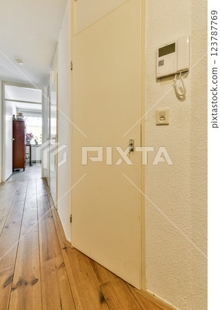 A spacious modern hallway features a wooden floor and light walls, leading to another room with a glimpse of furniture and natural light. A spacious modern hallway features a wooden floor and light walls, leading to another room with a glimpse of furniture and natural light. 123787769