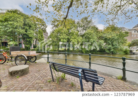 A serene view of a canal side with lush greenery, benches, and a bicycle cart enhancing the peaceful landscape, perfect for relaxation and nature appreciation. 123787969