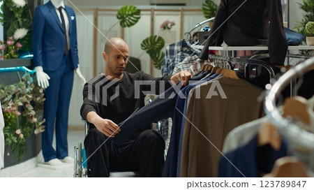 Middle eastern client in a wheelchair examining new clothes on racks at the retail store, consumerism and style choice. Young man with a chronic disability shopping for trendy items. Camera A. Middle eastern client in a wheelchair examining new clothes on racks at the retail store, consumerism and style choice. Young man with a chronic disability shopping for trendy items. Camera A. 123789847