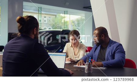 Customers in dealership completing necessary payment paperwork related to car sale, collaborating with finance and insurance departments to ensure smooth transaction, camera B Customers in dealership completing necessary payment paperwork related to car sale, collaborating with finance and insurance departments to ensure smooth transaction, camera B 123789968