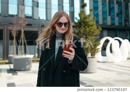 Smiling Caucasian blonde woman wearing trendy sunglasses walks down the central city street and uses her phone. Smiling Caucasian blonde woman wearing trendy sunglasses walks down the central city street and uses her phone. 123790197