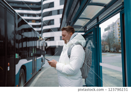 Young stylish man with smart phone on bus stop and typing sms stand at public transport bus station taking bus with headphones and mobile smart phone in winter or autumn day with backpack  123790231