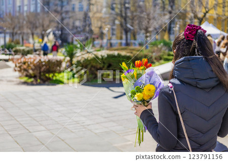 Young woman in black jacket holding bouquet of fresh colorful flowers in urban park, romantic mood. Sunlit spring atmosphere, blurred city background, romance. Vibrant springtime and celebration Young woman in black jacket holding bouquet of fresh colorful flowers in urban park, romantic mood. Sunlit spring atmosphere, blurred city background, romance. Vibrant springtime and celebration 123791156