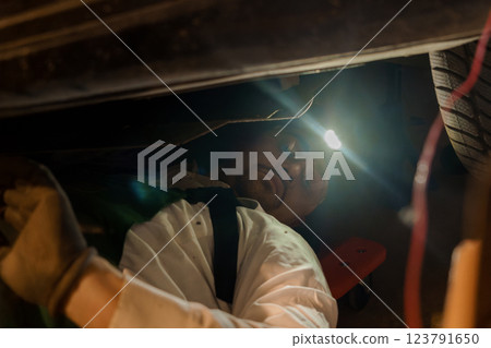 Mechanic in Gloves and Glasses Inspecting Vehicle Underneath Using Flashlight in Garage Mechanic in Gloves and Glasses Inspecting Vehicle Underneath Using Flashlight in Garage 123791650