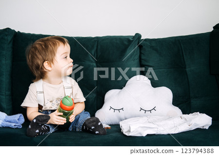 Side view of toddler holding sippy cup while sitting on couch with cloud pillow. Early childhood development, self-feeding, toddler independence, cozy home environment Side view of toddler holding sippy cup while sitting on couch with cloud pillow. Early childhood development, self-feeding, toddler independence, cozy home environment 123794388