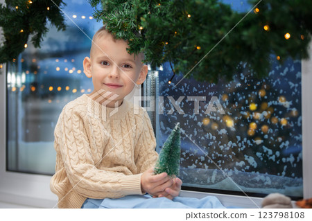 Little happy boy sits on the windowsill of a snow-covered window with Christmas tree decoration. Child in the Christmas room. Little happy boy sits on the windowsill of a snow-covered window with Christmas tree decoration. Child in the Christmas room. 123798108