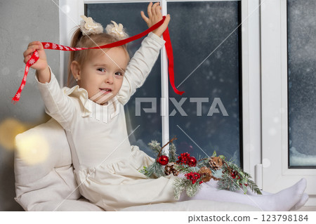 Little happy girl sitting on the windowsill playing with Christmas decor. 123798184