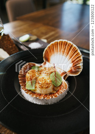 Grilled scallops in a shell with a crispy breadcrumb topping, adorned with fresh microgreens. Gourmet seafood appetizer presented on a black plate with a bed of salt 123800122