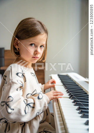 Home lesson on music for the girl on white piano synthesizer. Concept of early development: learning music enhances motor skills, hearing, and intelligence, fostering creativity from childhood Home lesson on music for the girl on white piano synthesizer. Concept of early development: learning music enhances motor skills, hearing, and intelligence, fostering creativity from childhood 123800895