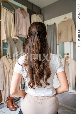 Young woman stylist in front of wardrobe filled with diverse clothing, analyzing outfit combinations. Creating fashionable looks requires attention to detail, color coordination, keen sense of style Young woman stylist in front of wardrobe filled with diverse clothing, analyzing outfit combinations. Creating fashionable looks requires attention to detail, color coordination, keen sense of style 123800898