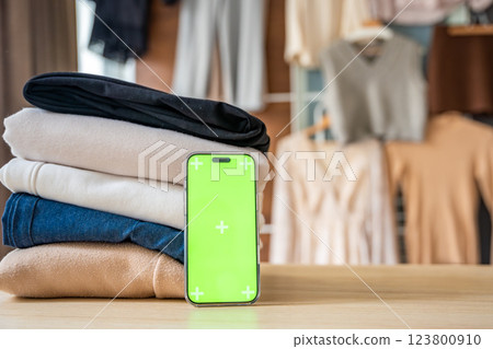 Stack of clothing prepared for resale with a smartphone displaying green screen for mockup use. Mobile apps simplify second-hand fashion, sustainable shopping, and conscious clothing redistribution 123800910