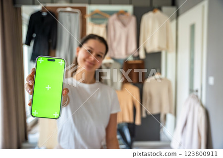 Young woman holds a smartphone close to the camera, displaying a green screen for mockup use. Mobile apps and digital tools transform online resale and second-hand fashion 123800911