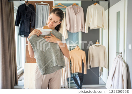 Young woman stands in front of a wardrobe filled with hanging outfits, holding clothing against herself. Choosing to wear is part of personal style, daily fashion decisions, and wardrobe organization 123800968