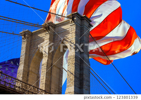 Magnificent view of Brooklyn Bridge featuring a giant American flag waving proudly against bright blue sky. 123801339