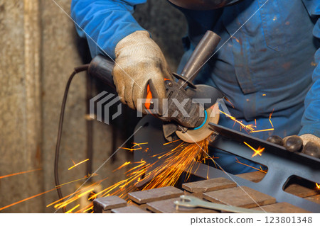 Metal worker uses grinder to smooth edges of steel while sparks illuminate dimly lit workshop. 123801348