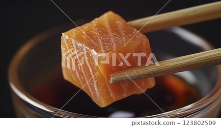 Fresh salmon cube held by chopsticks above soy sauce bowl in close-up 123802509