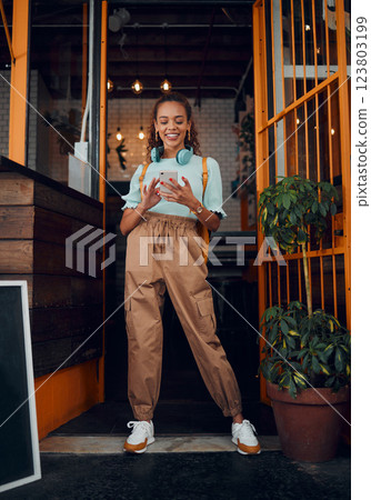 Cafe, student and phone social media with headphones on walk commute break in New York city. Happy urban college girl enjoying internet leisure on smartphone at doorway of local business. Cafe, student and phone social media with headphones on walk commute break in New York city. Happy urban college girl enjoying internet leisure on smartphone at doorway of local business. 123803199