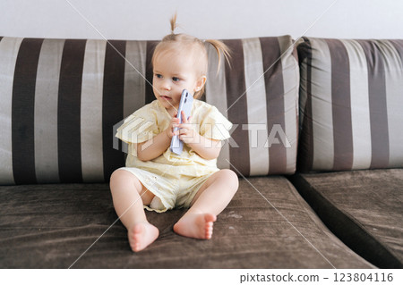 Portrait of child girl holding smartphone to ear, mimicking phone call. Joyful infant in light-colored outfit sitting on couch showcasing curiosity and imagination talking using smartphone at home. Portrait of child girl holding smartphone to ear, mimicking phone call. Joyful infant in light-colored outfit sitting on couch showcasing curiosity and imagination talking using smartphone at home. 123804116