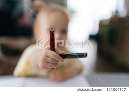 Blurred toddler girl showing two pencils to camera while sitting at table and drawing on paper at home, selective focus. Concept of childhood development, early learning, education. 123804127
