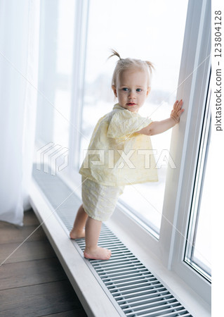 Vertical portrait of curious toddler girl with two playful ponytails standing by window on windowsill, soaking in winter scenery enjoying cozy warmth of home. Charming scene of childhood innocence. Vertical portrait of curious toddler girl with two playful ponytails standing by window on windowsill, soaking in winter scenery enjoying cozy warmth of home. Charming scene of childhood innocence. 123804128