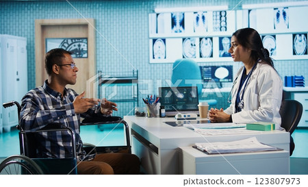 Indian general practitioner greeting wheelchair user patient to discuss illness with x ray results. Female medic doing a scan interpretation under medical insurance, health care. Camera B. Indian general practitioner greeting wheelchair user patient to discuss illness with x ray results. Female medic doing a scan interpretation under medical insurance, health care. Camera B. 123807973