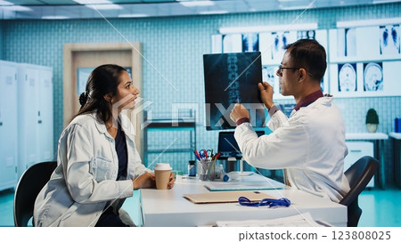 Professional medic and woman patient in a medical consultation looking at scans and MRI results, hospital records. General practitioner presents healthcare insurance to help with recovery. Camera B. Professional medic and woman patient in a medical consultation looking at scans and MRI results, hospital records. General practitioner presents healthcare insurance to help with recovery. Camera B. 123808025
