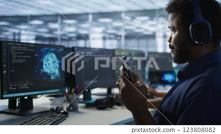 Worker in data center wearing headphones taking break from implementing AI systems, using smartphone. Focus on artificial intelligence server farm employee using mobile phone, camera A 123808082
