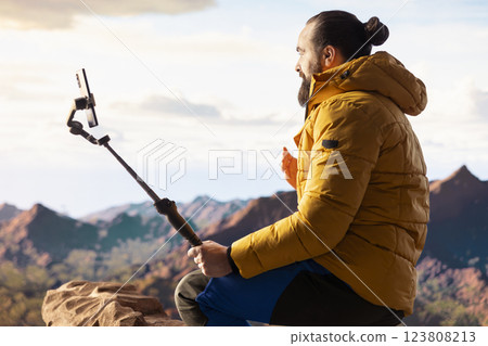 Content creator records a vlog in the mountains sharing his hiking journey with his online audience. Male explorer always connected with a smartphone mounted on a stabilizing tripod. 123808213