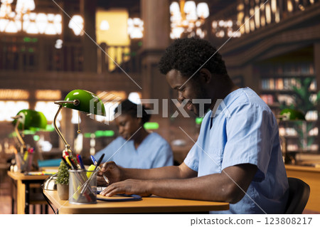 Black med student in scrubs does research and takes notes for his thesis, working hard at the campus library to become a doctor and obtain his diploma. Young man studying from copybooks. Black med student in scrubs does research and takes notes for his thesis, working hard at the campus library to become a doctor and obtain his diploma. Young man studying from copybooks. 123808217