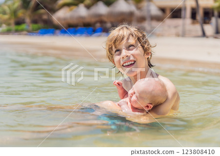 An older brother gently holds his baby sibling while swimming in the sea, sharing a joyful and heartwarming moment of family bonding and summer fun in the water. A perfect blend of love, care, and 123808410