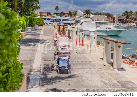 A young mother walks along the scenic waterfront, pushing a baby stroller with her child inside. A peaceful and heartwarming moment of motherhood, relaxation, and outdoor bonding by the sea 123808411