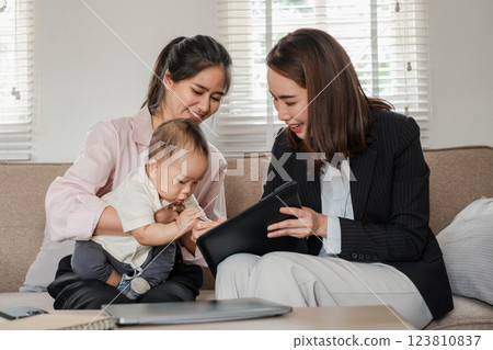 Two women and a baby in a home setting, discussing business with a tablet, showcasing work-life balance and modern family dynamics. Two women and a baby in a home setting, discussing business with a tablet, showcasing work-life balance and modern family dynamics. 123810837