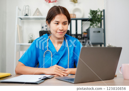 Young female doctor in blue scrubs using a laptop in a contemporary medical office setting, focused on work. 123810856