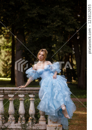 Romantic Woman in a Light Blue Gown Posing on a stone balustrade in a picturesque garden. 123812055