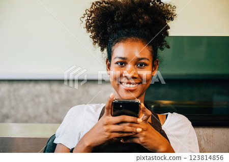 Close-up of a cheerful black barista coffee shop owner in uniform using a mobile phone. Portrait of the smiling woman working communicating joyfully. Entrepreneur's relaxation is evident. Close-up of a cheerful black barista coffee shop owner in uniform using a mobile phone. Portrait of the smiling woman working communicating joyfully. Entrepreneur's relaxation is evident. 123814856