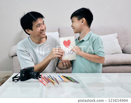 Smiling young boy presents handmade Father's Day card with red heart and "DAD" written on it to his father, sharing heartfelt moment at home 123814994