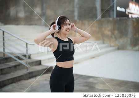 Joyful Asian young woman wearing wireless headphones while exercising outdoors in stylish athletic wear, enjoying music during her active lifestyle routine Joyful Asian young woman wearing wireless headphones while exercising outdoors in stylish athletic wear, enjoying music during her active lifestyle routine 123816072