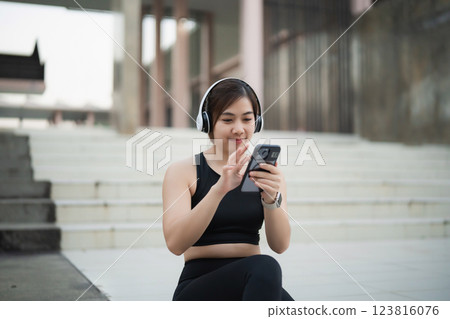 Asian Young woman in sportswear using smartphone and listening to music with headphones, sitting on steps in urban setting, enjoying a workout break 123816076