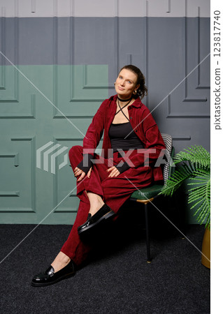 Cute woman in red shirt and loose trousers sits on chair by the colourful wall at home 123817740