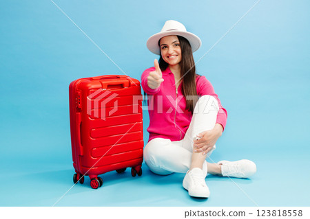 Happy young tourist woman sitting at the suitcase, showing finger up gesture and going to travel on holidays isolated on blue background. Happy young tourist woman sitting at the suitcase, showing finger up gesture and going to travel on holidays isolated on blue background. 123818558