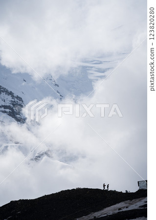 Vertical shot of two men working under huge snowy mountain shrouded with clouds, Pamir, Kyrgyzstan 123820280
