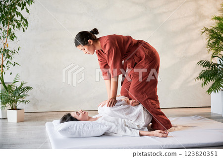 Masseuse making thai yoga massage. Woman therapist doing traditional massage treatment, stretching female patient's body. Client lying on mat, enjoying hand massage in calm atmosphere. 123820351