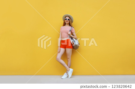 Beautiful young woman full length posing in summer hat with backpack on yellow 123820642