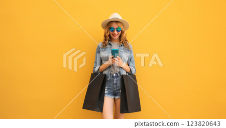 Beautiful happy young woman holding phone with black shopping bag, summer hat on yellow background Beautiful happy young woman holding phone with black shopping bag, summer hat on yellow background 123820643