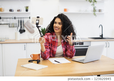 Young African woman wearing red flannel sits in kitchen recording video with smartphone. She expresses enthusiasm while gesturing, utilizing modern technology, and laptop on table. Young African woman wearing red flannel sits in kitchen recording video with smartphone. She expresses enthusiasm while gesturing, utilizing modern technology, and laptop on table. 123820945