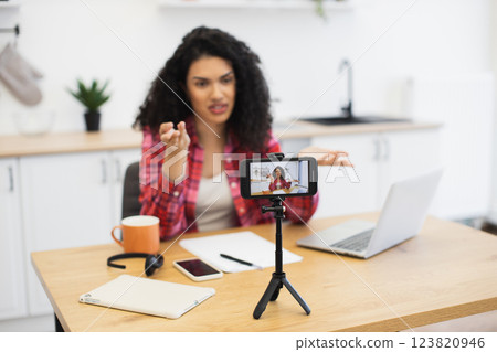 Young African woman talking to smartphone camera while working from kitchen table. Scene depicts modern technology usage, communication, remote work, and online content creation. Young African woman talking to smartphone camera while working from kitchen table. Scene depicts modern technology usage, communication, remote work, and online content creation. 123820946