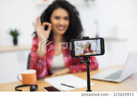 African woman in her 20s recording vlog using smartphone, wearing casual clothes, sitting in home office with coffee, papers, and laptop on table, creating engaging content for social media platforms. 123820953