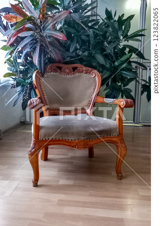 Vintage armchair with gray upholstery and wood carvings next to sprawling green bush in tub in room by window 123822065