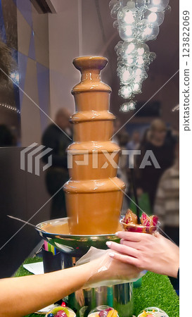 Salesman serves strawberries dipped in chocolate fountain to customer. Sale of sweet desserts. Close-up of hands 123822069